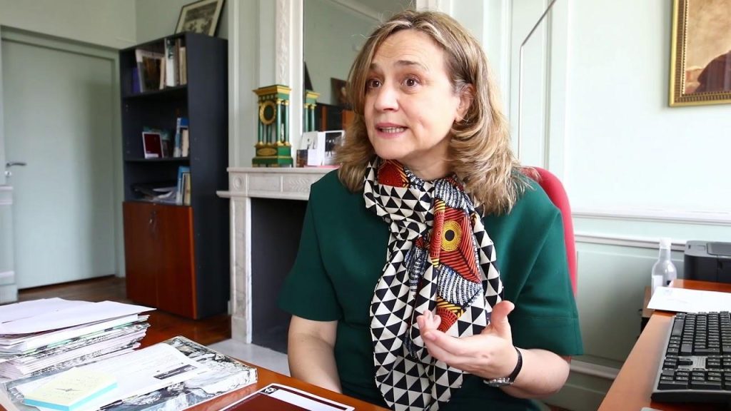 Luisa Castro durante entrevista, sentada ante escritorio con documentos, vistiendo cardigan verde sobre blusa estampada