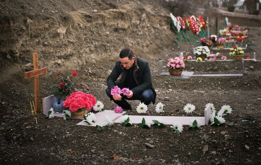 Persona de luto arrodillada ante una tumba con flores rojas en cementerio montañoso, en escena de duelo y remembranza