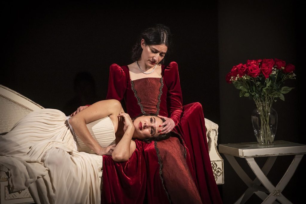 Mujer en vestido rojo de terciopelo reclinada en cama blanca junto a flores rojas, escena de drama lírico