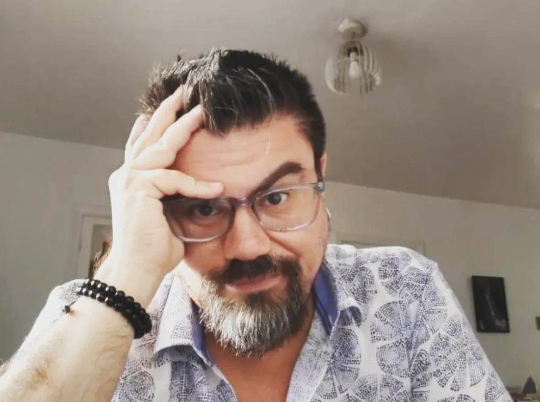 Hombre de barba oscura y gafas de marco rectangular, apoyando la cabeza en la mano derecha, en interior
