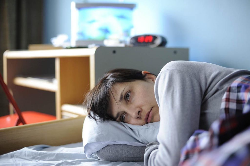 Mujer acostada en cama con expresión de tristeza y cansancio, habitación de fondo con muebles desenfocados