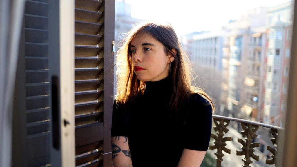 Mujer joven de cabello largo oscuro, vistiendo camiseta negra, en balcón con vista urbana al fondo