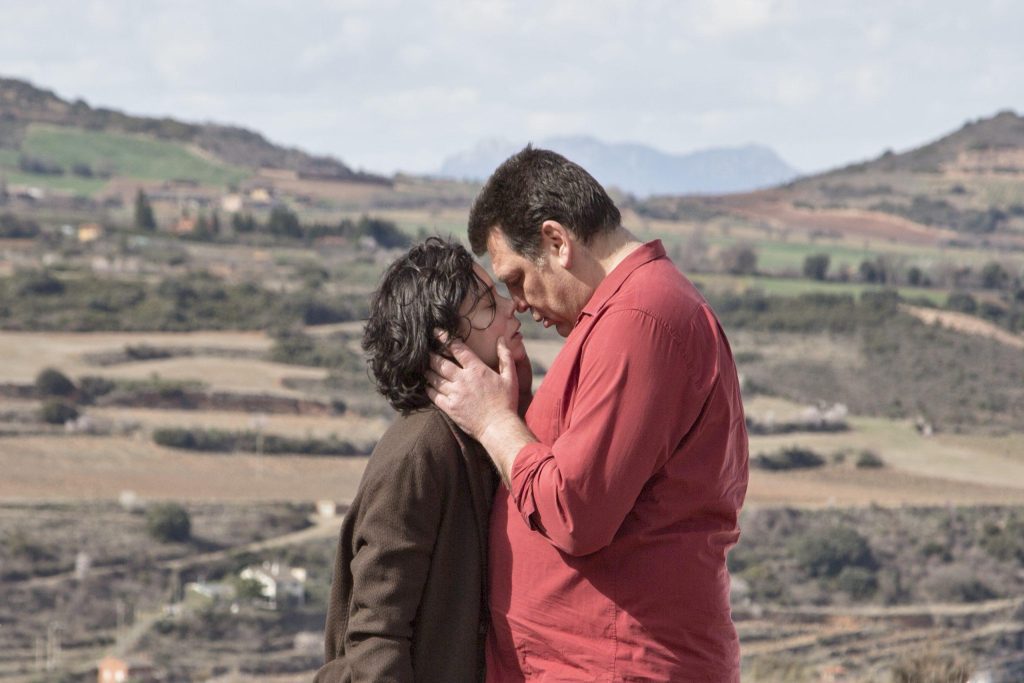 Pareja se besa en primer plano con paisaje rural montañoso de viñedos al fondo bajo cielo nublado