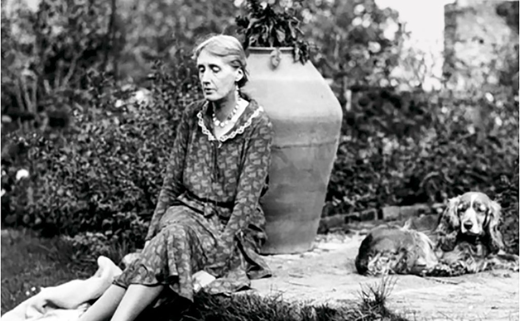 Virginia Woolf sentada en un jardín junto a una urna, con vegetación de fondo en fotografía en blanco y negro