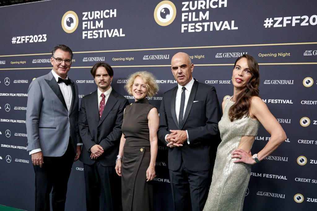 Cinco personas en traje de gala en alfombra roja del Festival de Cine de Zúrich frente a pared con logos