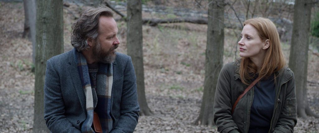 Dos personas sentadas conversando en un bosque entre árboles, en una escena de diálogo íntimo y reflexivo