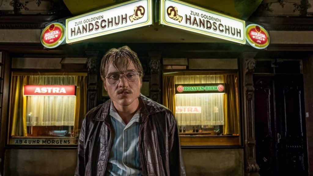 Hombre con gafas y bigote frente a fachada iluminada de Handschuh, tienda histórica de Hamburgo
