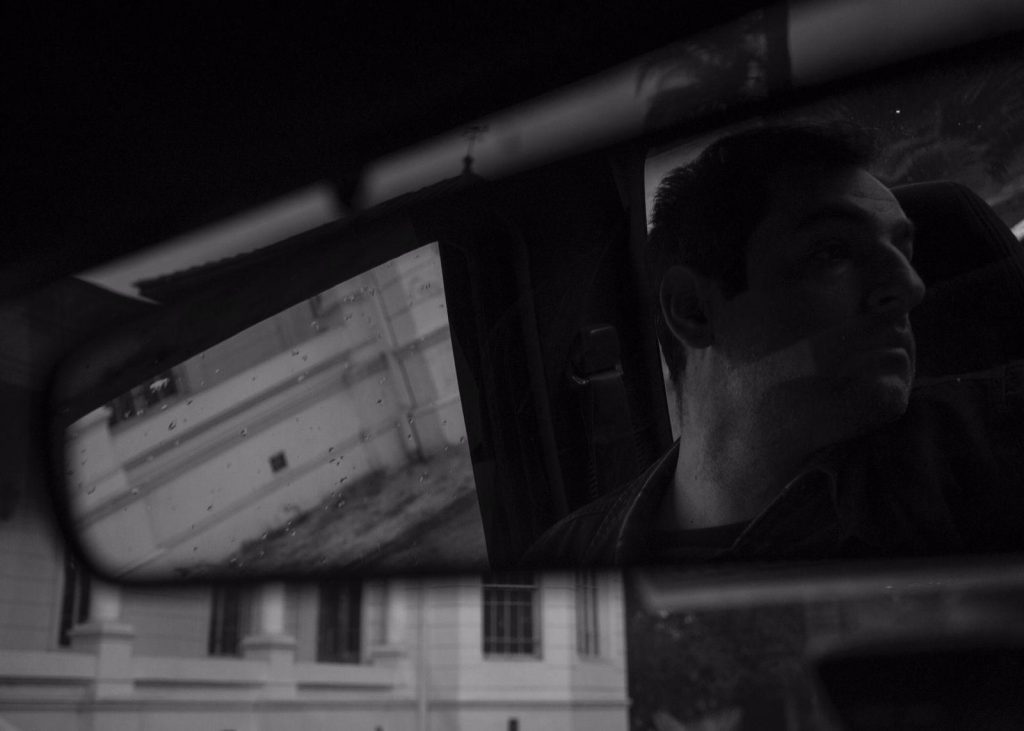 Hombre de perfil en automóvil nocturno, pantalla de edificio reflejada, atmósfera noir en blanco y negro