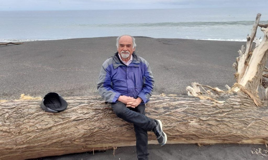 Juan Mihovilovich sentado sobre tronco de madera junto a acantilado y playa, vistiendo chaqueta azul