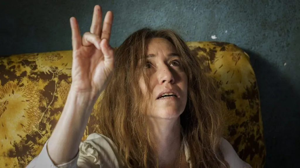 Mujer con cabello largo y ondulado levanta la mano en gesto de paz, sentada frente a fondo amarillo texturizado