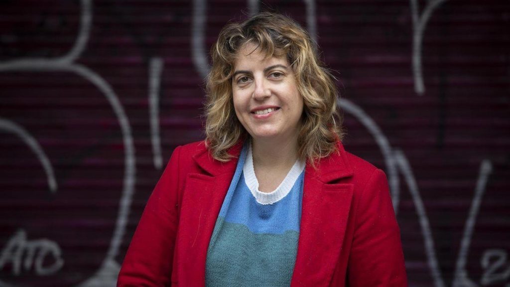 Mujer sonriente con ondas rubias, blazer rojo y camiseta azul sobre fondo con graffiti gris