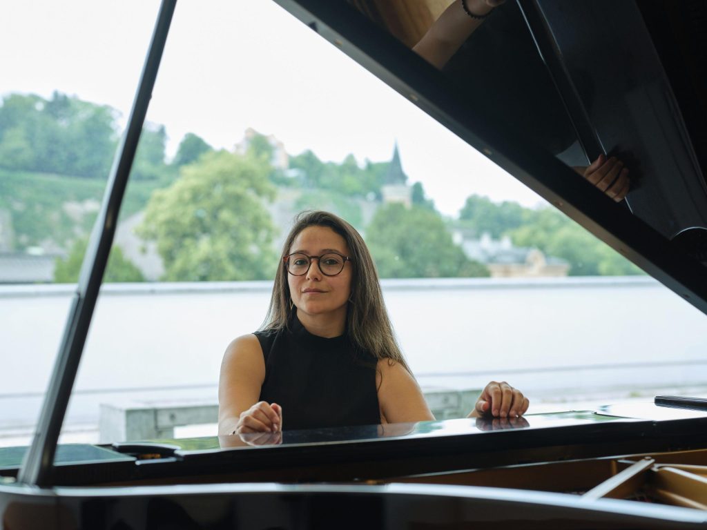 Montserrat Bravo al piano frente a un lago con árboles y casas al fondo