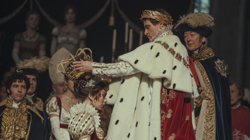 Escena de coronación imperial: Napoleón Bonaparte viste uniforme blanco y capa de armiño mientras personajes
