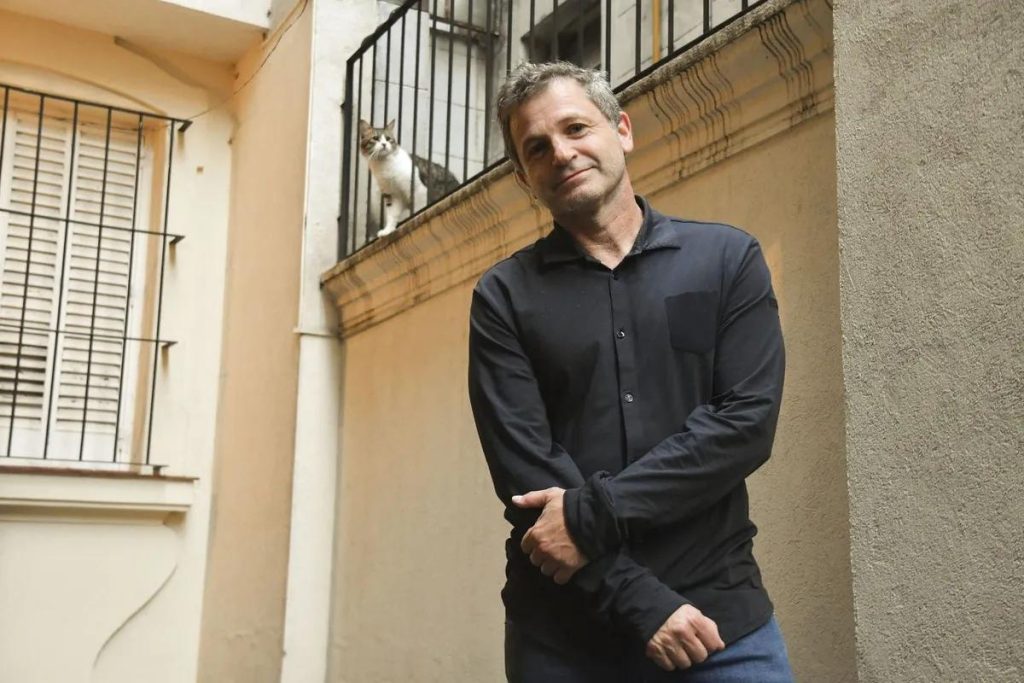 Hombre de mediana edad en chamarra negra, parado en pasillo de arquitectura colonial con escaleras metálicas al fondo