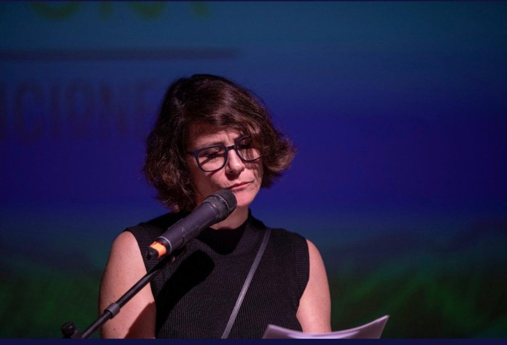 Sara Bertrand con micrófono en evento cultural nocturno, iluminación azul de fondo, expresión concentrada