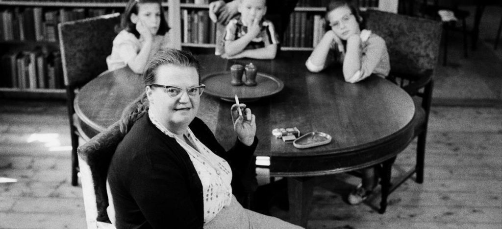 Shirley Jackson sentada en una silla, con gafas, en un interior de mediados del siglo XX con libros al fondo