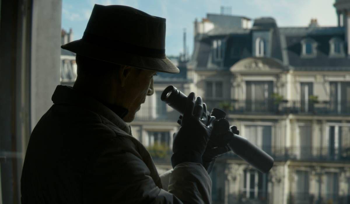 Hombre de sombrero y abrigo oscuro observa desde una ventana parisina con binoculares, ciudad de fondo