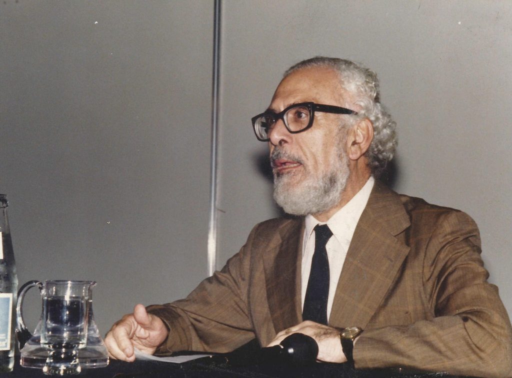 Antonio Di Benedetto, escritor argentino con gafas oscuras, sentado ante micrófono en panel de conferencia