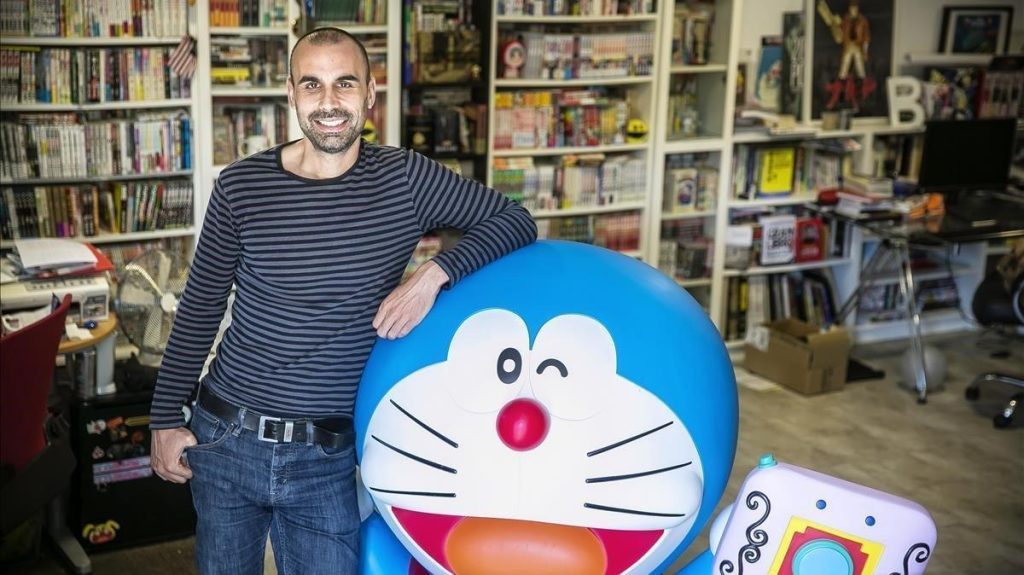 Marc Bernabé junto a figura de Doraemon en biblioteca con estanterías de libros y películas al fondo