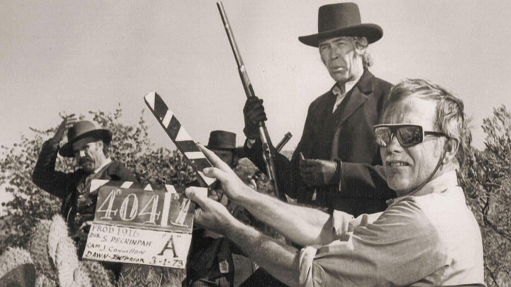Sam Peckinpah dirigiendo una escena de acción, sosteniendo una cámara de cine en exteriores junto a su equipo