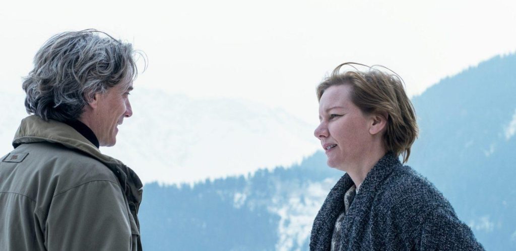 Dos personas conversan en un bosque nevado con montañas de fondo, escena de tensión dramática en ambiente invernal