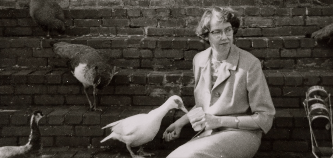 Flannery O'Connor sentada con patos en sus brazos frente a pared de ladrillos