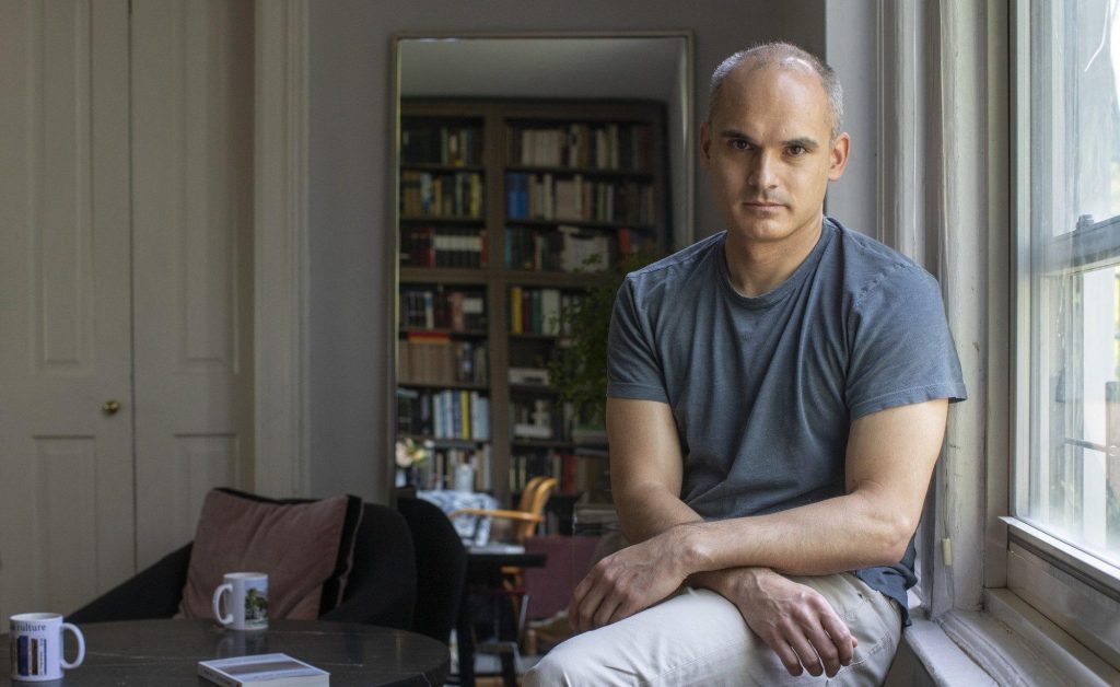 Hombre de mediana edad sentado en escritorio junto a ventana, con estantería de libros al fondo, mirando a cámara