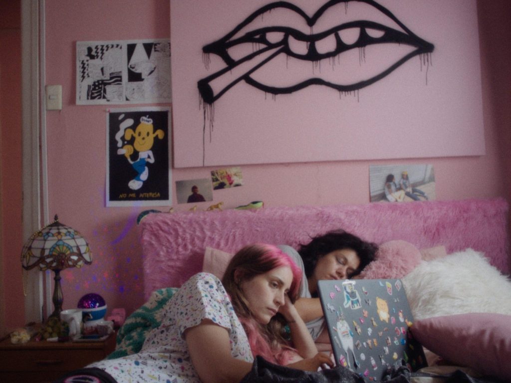 Habitación adolescente con paredes rosas, dos chicas en cama con almohadas de colores, carteles en pared, decoración
