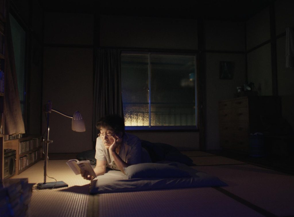Hombre sentado en habitación oscura iluminada por luz de ciudad nocturna, mirando hacia la ventana con expresión