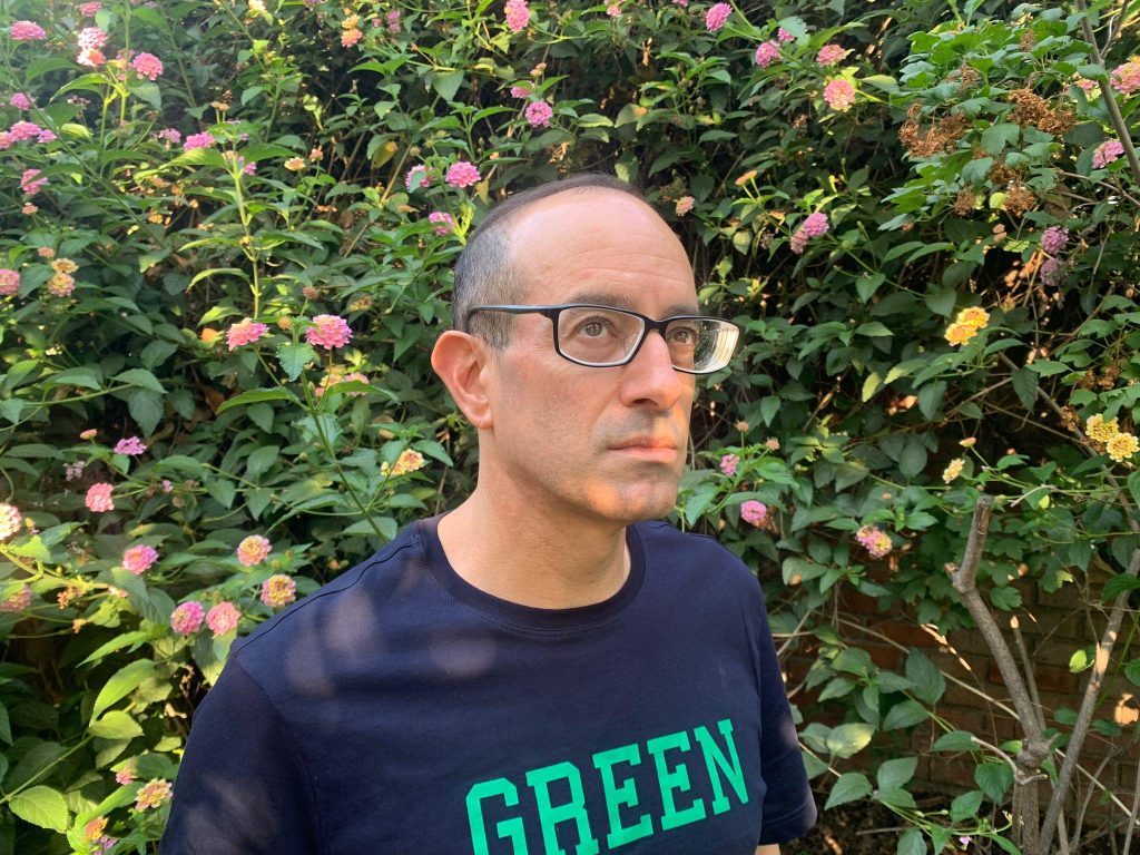Nicolás Poblete, escritor con lentes y polera negra con texto verde, frente a arbustos con flores rosas
