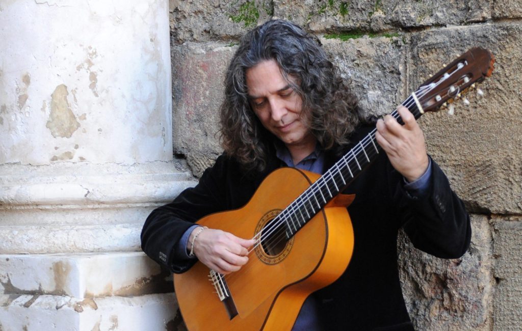 Tomatito sostiene su guitarra clásica mientras posa contra una pared de piedra gastada, vistiendo chaqueta negra