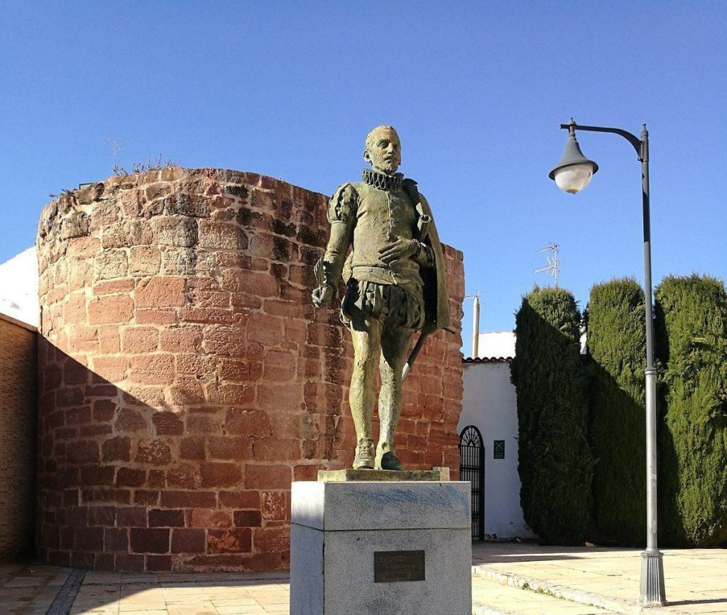 Estatua de Miguel de Cervantes Saavedra en piedra roja, junto a muralla antigua y farola bajo cielo despejado