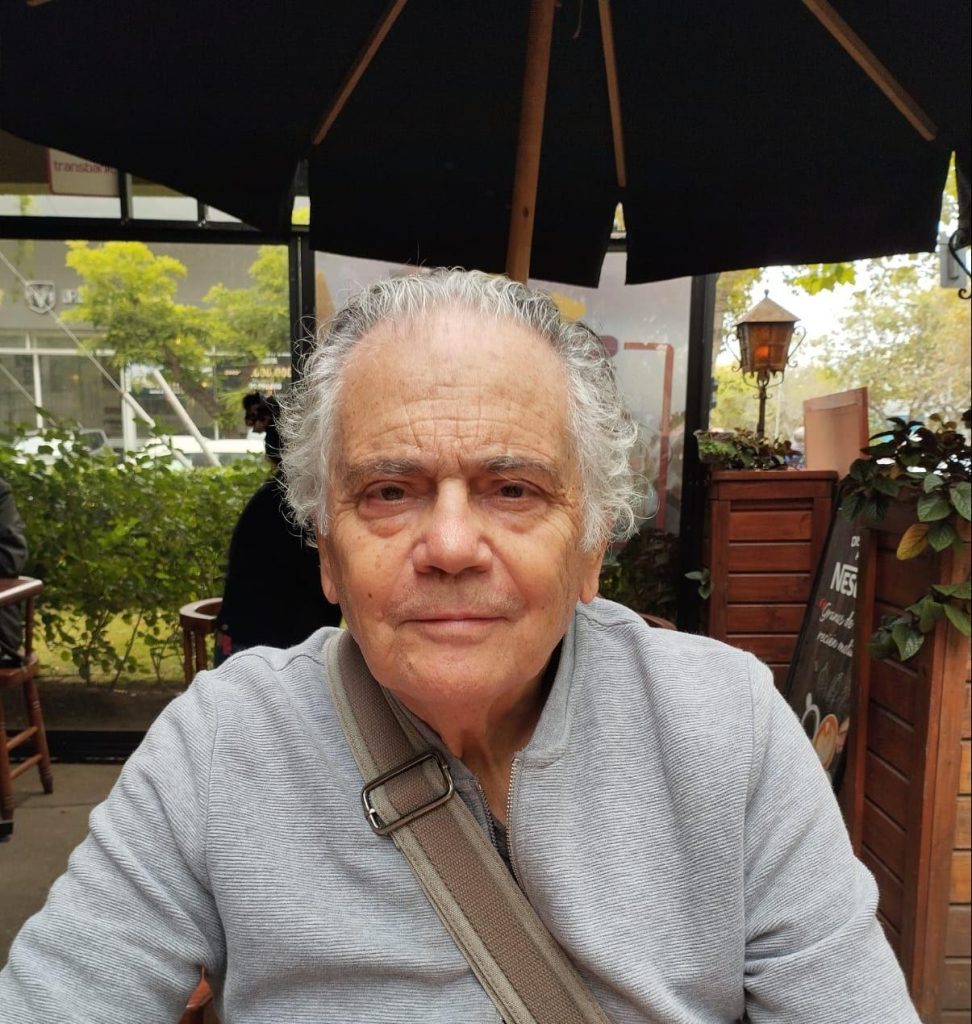 Edmundo Moure, escritor y académico, bajo sombrilla en jardín, mirando a cámara con expresión serena