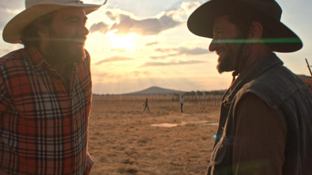 Dos cowboys conversan al atardecer en un árido paisaje desértico bajo un cielo nublado con tonos dorados