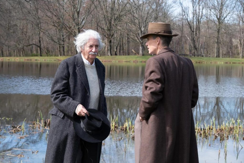 Dos hombres conversan junto a un lago en escena de película, uno con abrigo oscuro y otro con sombrero de fieltro