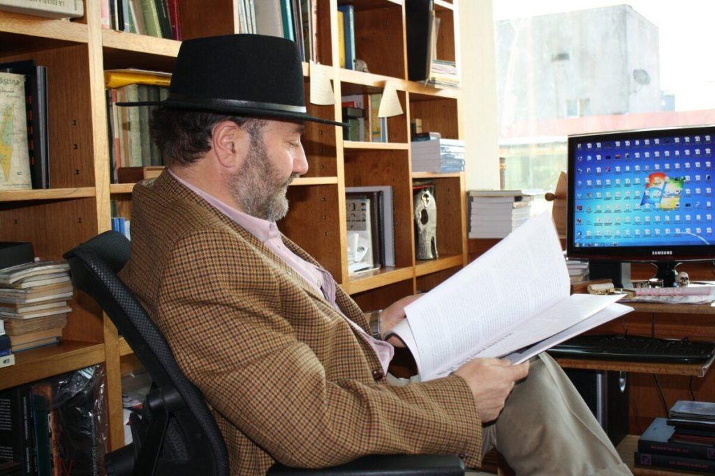 Theodoro con sombrero negro y abrigo marrón sostiene papeles en su escritorio, frente a una computadora