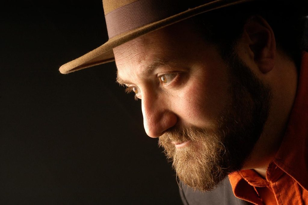 Theodoro Elssaca, hombre de barba con sombrero y camisa naranja, retrato de perfil sobre fondo oscuro