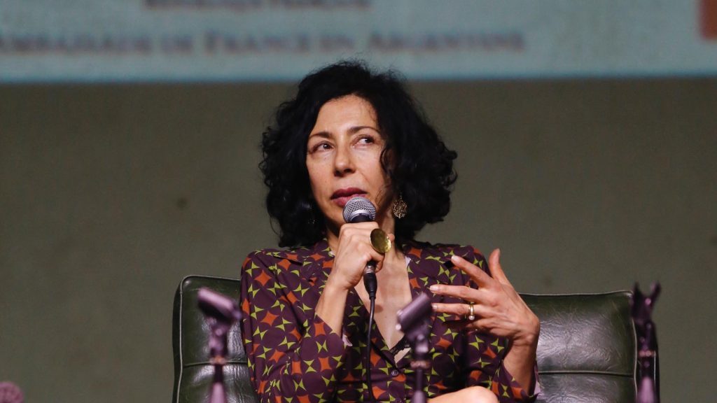Yasmina Reza durante una conferencia, gesticulando mientras habla, vistiendo blusa estampada sobre fondo neutro