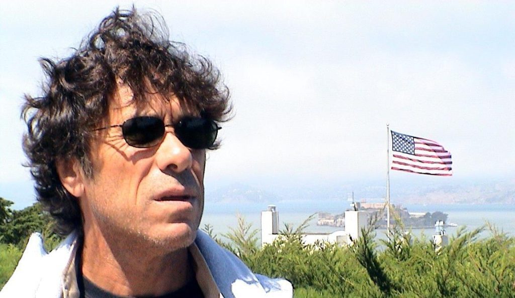 Emilio Pacull con gafas de sol, camisa blanca y paisaje urbano con bandera estadounidense al fondo