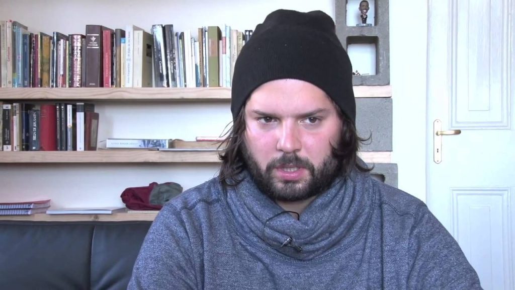 Hombre con gorro negro y buzo gris oscuro, barba poblada, mirando a cámara en interior con estantería de libros al fondo