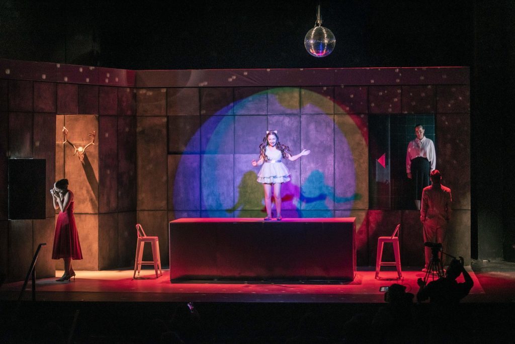 Escena teatral con actriz en centro iluminado de azul, flanqueada por dos figuras en rojo oscuro sobre escenario