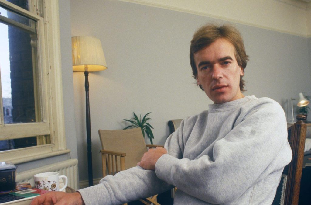 Martin Amis en interior moderno, sentado en silla de madera, viste suéter gris claro y mira hacia la cámara