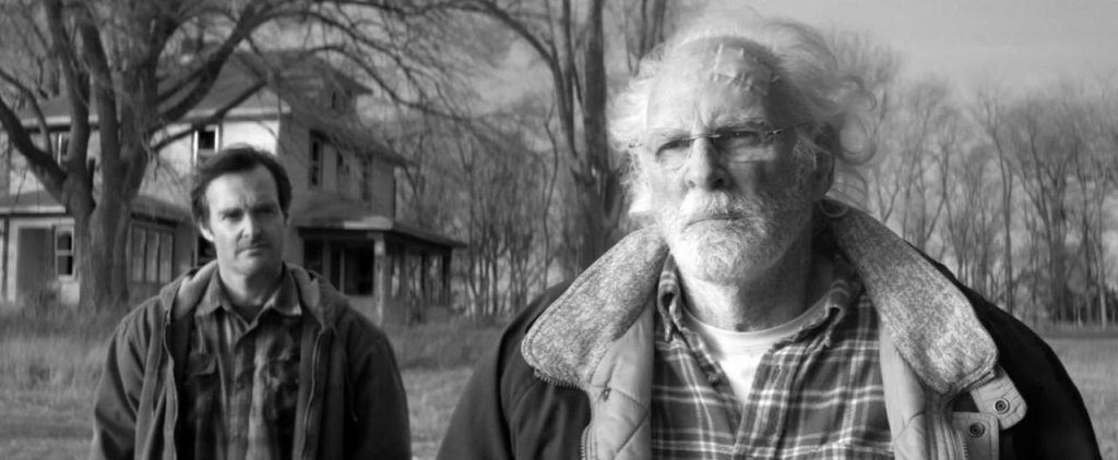 Dos hombres en camino rural de Nebraska: un anciano con chaqueta de cuero y un joven, árboles desnudos al fondo