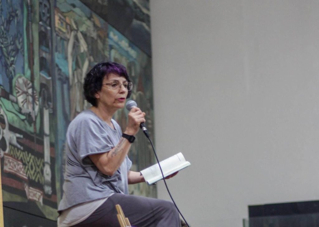 Silvia Moreno-Garcia sostiene micrófono durante presentación en evento cultural, con pantalla de proyección al fondo