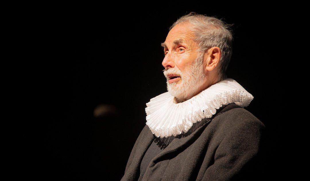 Hombre de mediana edad con cabello gris, perfil izquierdo, viste jubón oscuro con cuello blanco voluminoso, fondo negro