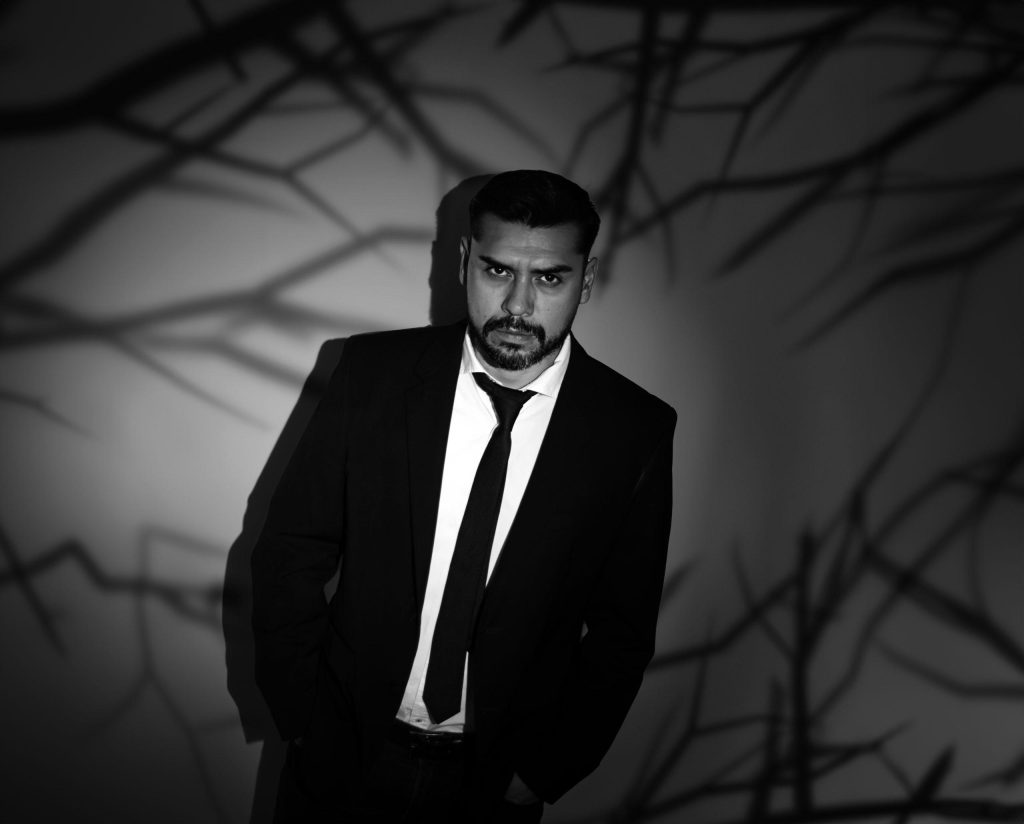 Retrato en blanco y negro de Jesús Diamantino con traje y corbata, sobre fondo con ramas abstractas