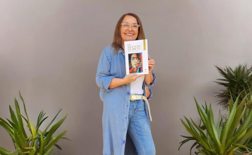 Mujer sostiene libro titulado El silencio de Irene junto a plantas verdes, viste camisa azul clara