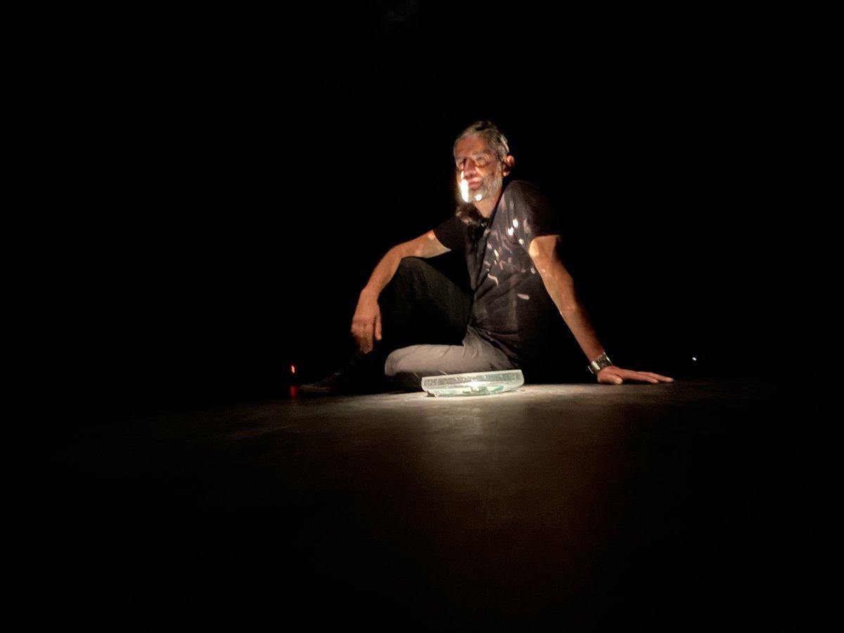 Hombre de mediana edad sentado ante una mesa iluminada, con expresión intensa y contemplativa en ambiente teatral oscuro
