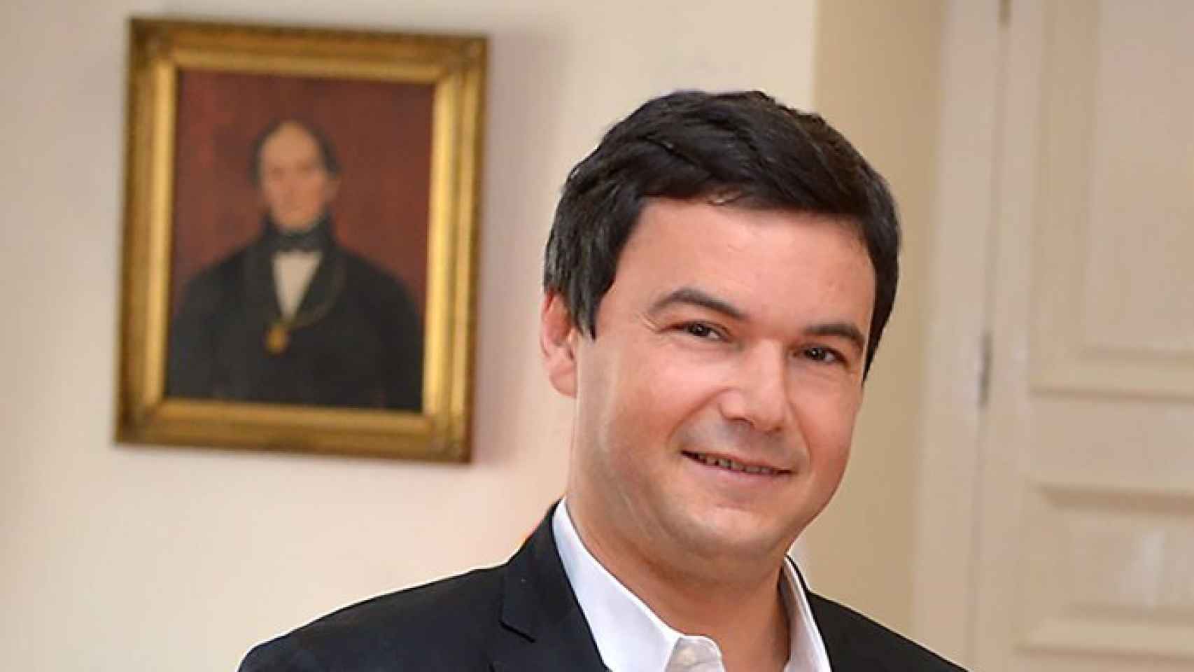 Hombre de mediana edad con traje oscuro sonriendo ante un retrato al óleo, en interior elegante