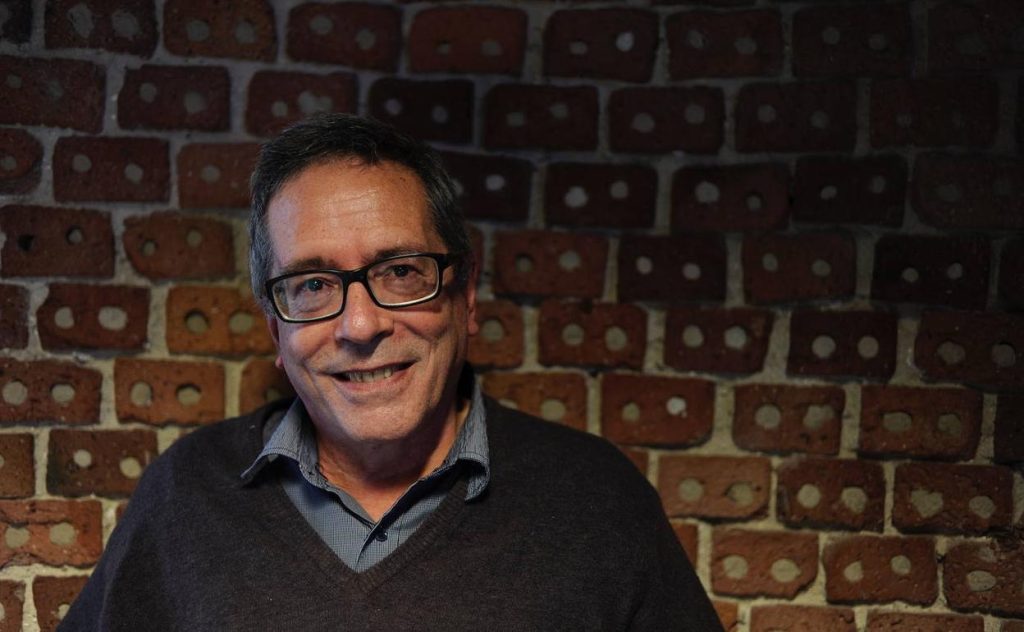Hombre con gafas y suéter oscuro frente a pared de ladrillos rojos, sonriendo a la cámara